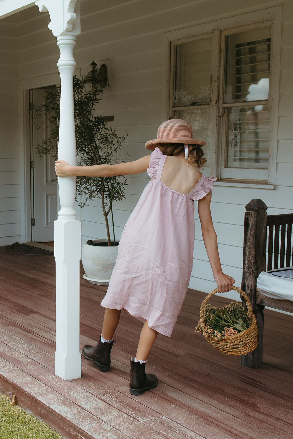 Young girl in a pink linen dress and hat tending to flowers in a garden with a white arbor.