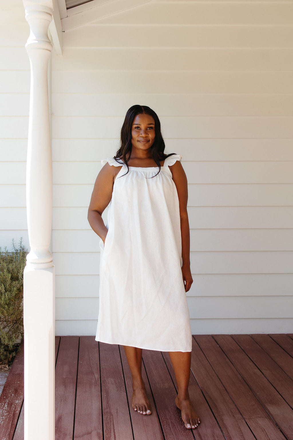 Woman in a white linen nightie standing on a wooden deck with a white wall background