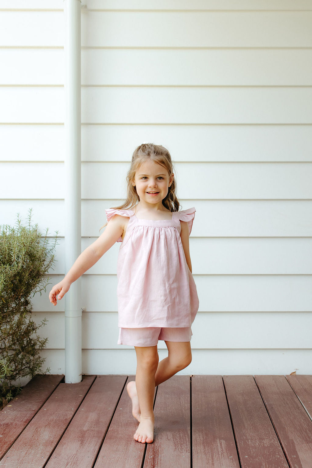 Young girl in a pink linen shorts and top standing on a wooden deck against a white wall.