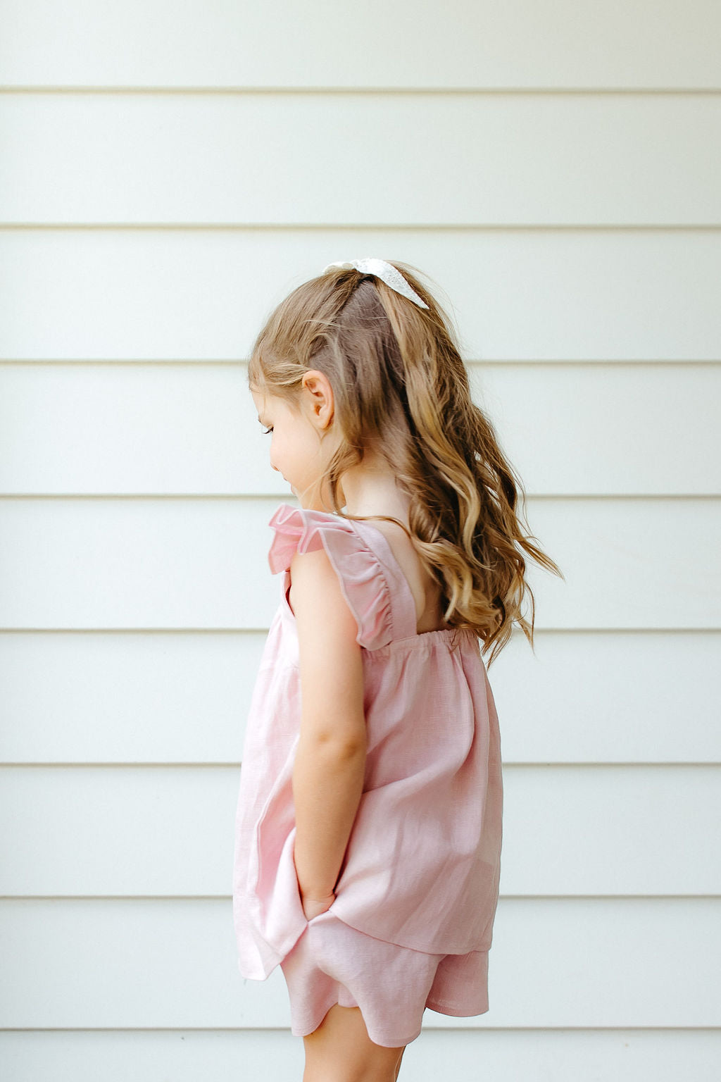 Young girl in a pink linen shorts and top standing on a wooden deck against a white wall.