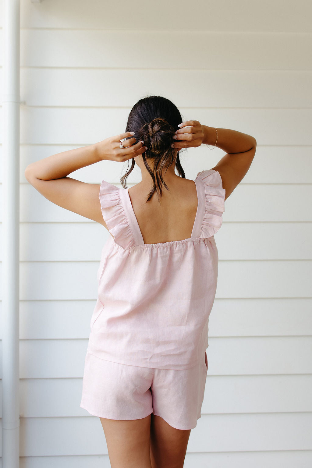 Woman wearing a pink linen top and shorts with ruffled straps against a white wall.